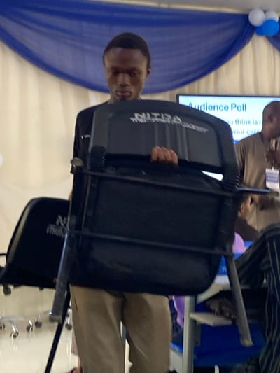 A young man with short dark hair, wearing a black shirt and light brown pants, stands centrally in an indoor event space in Baganakwo, Nigeria. He holds a black folding chair vertically in front of him, partially obscuring his face and torso; his left hand is visible gripping the chair's top frame. The chair's backrest features white text, "NITDA THURSDAY." In the background, a large screen displays "Audience Poll" with additional obscured text, indicating a question. To the right of the screen, a man in a tan long-sleeved shirt with an ID badge is partially visible. The room's backdrop consists of a white wall adorned with blue draped fabric and white and blue balloons. Another black chair, similar in design, is visible to the left of the primary subject. The environment suggests a formal gathering or conference setting.