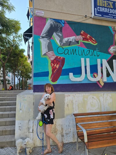 A woman is holding a small white dog while standing next to a colorful mural on a building. The mural depicts a stylized figure with red shoes and striped socks. The text "Caminant" and "JUN" is visible on the mural. To the left of the mural, a blue sign with white text reads "IBEX CORREDOP AUTOMÒ COMERÇOS CO". The woman is wearing a floral dress and sandals, and she is holding the dog in her arms. Another small white dog is visible on the ground to her right, on a blue leash. In the background, there are trees, buildings, and a clear sky, suggesting it is daytime. The overall scene appears to be outdoors, possibly in a public space like a street or park. The woman's expression is neutral, and she is looking towards the camera. The setting seems to be warm and sunny.