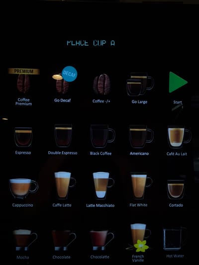 This image shows a digital display of a coffee machine menu. The display is black with white and blue text and images. At the top, the text "PLACE CUP A" is visible. Below this, there are several coffee options presented with corresponding images. From left to right and top to bottom, the options are:
Top row:
- "PREMIUM" text above a coffee bean image, labeled "Coffee Premium"
- A light shining on a coffee bean image, labeled "DECAF" and "Go Decaf"
- A coffee bean image, labeled "Coffee -/+""
- Two stacked coffee cups, labeled "Go Large"
- A green play button, labeled "Start"
Second row:
- A glass cup with dark liquid, labeled "Espresso"
- A glass cup with dark liquid, labeled "Double Espresso"
- A glass cup with dark liquid, labeled "Black Coffee"
- A glass cup with dark liquid, labeled "Americano"
- A glass cup with light brown liquid, labeled "Café Au Lait"
Third row:
- A glass cup with light brown liquid and foam, labeled "Cappuccino"
- A tall glass cup with light brown liquid and foam, labeled "Caffe Latte"
- A tall glass cup with layered light brown liquid and foam, labeled "Latte Macchiato"
- A glass cup with light brown liquid, labeled "Flat White"
- A glass cup with dark liquid and foam, labeled "Cortado"
Fourth row:
- A cup with dark liquid and foam, labeled "Mocha"
- A cup with dark liquid, labeled "Chocolate"
- A cup with dark liquid and foam, labeled "Chocolatte"
- A glass with a yellow flower and a cup with light brown liquid, labeled "French Vanille"
- A glass cup filled with clear liquid, labeled "Hot Water"
The image appears to be taken indoors, likely in a commercial setting where a coffee machine is present. There is no information in the image that allows for determination of its location in the Town of East Hampton, United States of America. The scene is a digital interface on a screen, with no people or specific environmental details visible beyond the display itself.