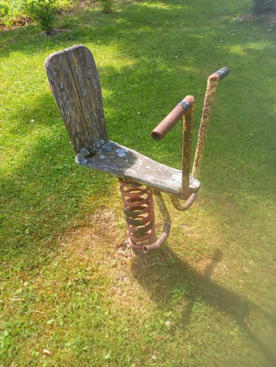 An aged, wooden-seated spring rider, weathered and showing signs of lichen growth, is situated on a patchy lawn with areas of dry grass. The metal structure supporting the seat and handlebars is rusty, painted yellow and red, with some black tape on the handlebars. The rider is positioned in a grassy area, with shadows from nearby foliage cast across the lawn. The overall scene suggests an outdoor recreational space, possibly a park or a backyard, located in Adutiškis, Lithuania.