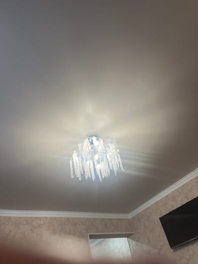 The image shows an interior scene, likely a room. The ceiling is visible, featuring a modern chandelier with numerous clear, tear-drop-shaped crystals hanging from a metallic base. The chandelier is illuminated, casting light and shadows on the ceiling. Below the ceiling, a white decorative cornice runs along the wall. The wall itself is covered with textured wallpaper that has a marble-like pattern in shades of beige and brown. To the right side of the image, a portion of a black flat-screen television is mounted on the wall. An open doorway or alcove is partially visible on the left side of the wall. The lighting suggests an indoor environment. There is no indication of the specific location in Nalchik, Russia, or any people in the image.