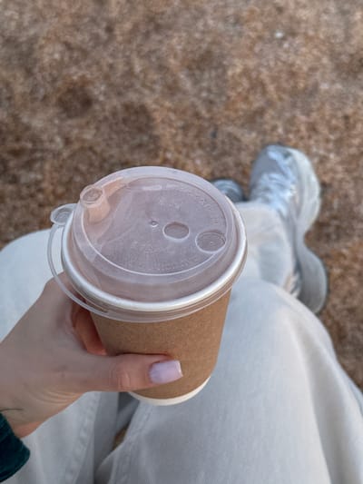 A top-down, medium shot depicts a person holding a brown paper coffee cup with a clear plastic lid. The lid has a drinking spout and air holes. The person's hand, with a pale purple fingernail, is visible. The person is wearing light-colored trousers and their shoes are slightly visible in the background. The ground appears to be a textured, brownish surface, possibly sand or concrete. The location is not possible to discern from the image.