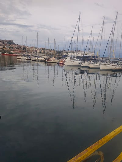 The image depicts a harbor in Kavala, Greece, with numerous sailboats docked. The city's characteristic colorful buildings and a fortress are visible on the hillside in the background. A large ferry is docked behind the sailboats. The water in the foreground is calm, reflecting the masts of the boats and the cloudy sky. The overall scene is tranquil, suggesting a peaceful maritime setting in the port of Kavala.
