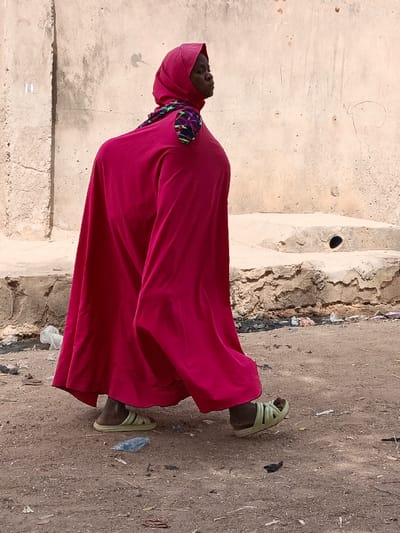 An individual is depicted walking on a dirt path in Zango, Nigeria. The subject, appearing to be female, is dressed in a full-length, bright magenta garment that covers the body from head to ankle, including a matching head covering. A multicolored patterned fabric, featuring geometric shapes in green, blue, purple, and black, is visible draped over the left shoulder. Light green, open-toed slip-on sandals are worn on the feet. The individual's body is oriented towards the right of the frame, but the head is turned to the right, showing the right profile of the face. The ground is unpaved, consisting of dry, reddish-brown dirt with scattered debris, including fragments of white and blue plastic. In the background, a light beige wall, exhibiting an uneven, weathered texture, rises from a rough concrete or stone ledge. A dark, circular hole is present in this concrete ledge on the right side of the frame.