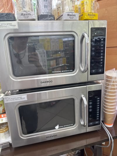 The image displays two stacked Daewoo "PROFESSIONAL MICROWAVE OVEN" units, constructed from stainless steel, situated on a dark brown counter or table. The setting is an indoor commercial space, consistent with a convenience store or similar establishment in South Korea, as evidenced by Korean text on product packaging and appliance labels. The precise location within 종로구 is not discernible from the image.
The upper microwave features an active digital display showing "0" and a control panel with various time settings (e.g., 30 seconds, 1, 2, 3, 5, 10, 30, 40 minutes) and Korean function buttons for "Cancel" and "Start." Reflected in its glass door is a person, likely the photographer, wearing a white shirt and a dark tie or neck accessory, holding a smartphone. Above this microwave, several plastic-wrapped retail products, including boxes of "Maxim White Gold Coffee Mix," are visible.
The lower microwave is visually similar to the upper unit, with its display off. A small, irregular white mark or tear is present on its door window. A white sticker with Korean text, stating "Movement occurring after cooking completion is not a malfunction, so use with peace of mind," is affixed to its upper left corner. To the right of the lower microwave, a transparent plastic bag contains a stack of light brown, disposable paper cups. The background, reflected in the microwave doors, shows shelves stocked with various packaged goods, typical of a retail environment. The wall behind the counter appears to be light-colored wood paneling.