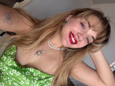 A young woman with blonde hair is lying down and looking at the camera with a smile. She is wearing a green dress with white flowers and a pearl necklace with a pendant. She is also wearing earrings. A butterfly tattoo is visible on her arm. The background appears to be a room with a white wall and a glimpse of a window or mirror. There is no information to indicate the location is in the city of Большая Шильна, Russia.