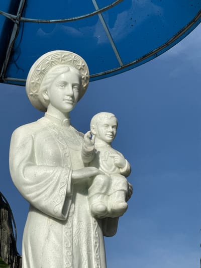 A white marble statue of Mary holding the infant Jesus stands against a bright blue sky. The statue is positioned under a blue, circular canopy supported by metal framing. Mary wears a hat adorned with stars and a traditional Vietnamese dress with intricate embroidered patterns on the collar and sleeves. Jesus is depicted as a young child held in her arms, with his right hand raised in a blessing gesture. The statue exhibits fine detail in the sculpting of the facial features and drapery. The background is a clear, cloudless blue sky, with hints of dark foliage and tree trunks visible on the left edge of the frame. The overall scene appears to be outdoors, likely in a public space or religious site within the city of Đa Thọ, Vietnam.