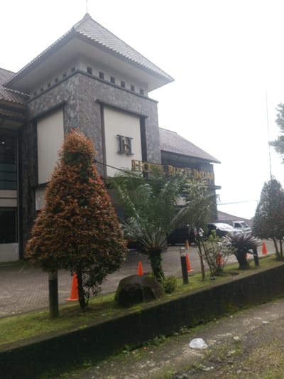 The image presents the exterior of the "HOTEL BUKIT INDAH," located in Thanthirimale, Sri Lanka. The multi-story building features a brown tiled roof and a stone-clad central facade with lighter, rectangular panels. A large, stylized "B" emblem is affixed to one panel, while golden lettering spelling "HOTEL BUKIT INDAH" is displayed below it. In front of the hotel, a paved area is partially visible, containing several parked vehicles, including a dark SUV and a white van. The foreground features a landscaped garden bed bordered by a low concrete wall. This bed contains a prominent conical tree with dense reddish-brown foliage, several smaller palm-like plants, and a large dark rock. Multiple orange traffic cones are distributed within the garden, alongside two dark, cylindrical bollard lights. The immediate foreground shows a dirt patch with sparse grass and a crumpled white plastic bag. The sky is uniformly overcast. No individuals are visible within the scene.