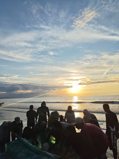 A group of fishermen are gathered on the beach at Uleekareung, Indonesia, during sunset. The sky is a mix of blue with scattered white clouds, and the sun is setting over the ocean, casting a golden glow on the water. The fishermen are silhouetted against the bright sky, and they appear to be working with fishing nets and a catch of fish. Some of the fishermen are wearing headscarves or hats. The waves are gently lapping at the shore.