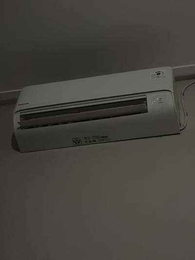 A white Panasonic air conditioner is mounted on a light gray wall, likely in a residential or office setting. The air conditioner has a light gray front panel with horizontal louvers that are slightly open, suggesting it is either in operation or recently powered off. There are indicator lights on the right side of the unit, with a green light illuminated, possibly indicating power or operation. A cable is connected to the right side of the air conditioner and runs down the wall. The wall appears to be painted in a matte finish. The lighting in the room is dim, creating shadows and highlighting the textures of the wall and the air conditioner. There are no people visible in the image. The scene does not provide any specific information about its location within Tupah, Indonesia, beyond being an indoor environment.
