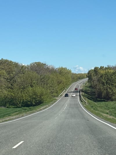 A view from above depicts a grey asphalt road curving through a rural landscape under a clear blue sky. Lush green trees and grass line the sides of the road. Several vehicles are seen driving on the road. The location appears to be Pokrovskoye, Russia, based on details like the type of foliage and the style of road.