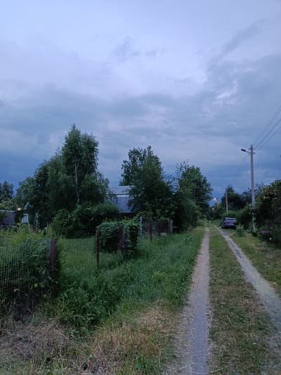 A dirt road runs through a rural area in Alferovo, Russia, with dense foliage lining both sides. The sky is overcast with dramatic, dark clouds. On the left side of the road, a wire fence is partially visible, behind which a house with a dark roof and a window can be seen. Further down the road, a dark-colored car is parked. Utility poles with wires are also present, suggesting the presence of electricity. The overall scene depicts a somewhat gloomy, late afternoon or early evening setting in a countryside environment.