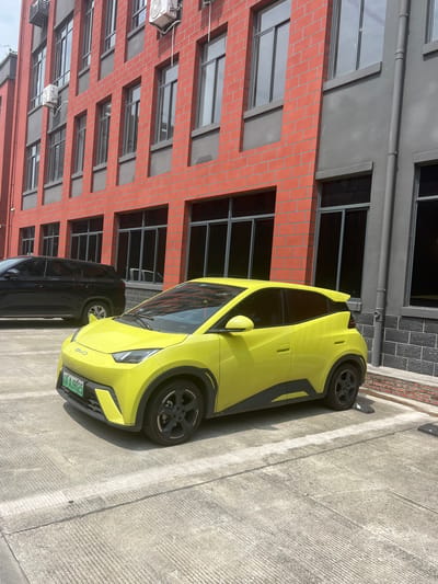 A bright yellow compact electric car is parked on a concrete surface in front of a red brick building. The car has tinted windows and black accents around the wheel wells and along the lower body panels. A license plate with green lettering is visible on the front of the car. To the left of the yellow car, a dark-colored SUV is partially visible. The building behind the cars features rows of windows and appears to be an office or commercial building. The environment suggests an urban setting in Shuangfu, China, possibly a parking lot associated with the building. The lighting indicates it is daytime.