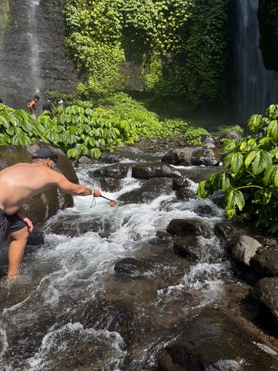 A man wearing black shorts and a black baseball cap stands in a shallow, rocky river, facing left. He is shirtless and has tattoos on his left arm. He holds a selfie stick with an orange object attached to the end, extending it towards the water. The river flows with white foam over dark, wet rocks. In the background, lush green vegetation covers a dark cliff face, with a waterfall cascading down from the top left of the frame. Two other people, one shirtless and one wearing a black shirt, are visible near the waterfall in the background. The scene appears to be at a natural waterfall in Sawan, Indonesia, a region known for its scenic beauty and waterfalls.