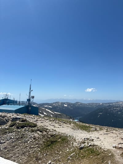 The image depicts a high-altitude landscape, likely a mountaintop, with a clear blue sky dominating the upper portion. In the mid-ground, a blue building with a corrugated metal roof is visible, accompanied by a tall communications tower featuring multiple antennas and equipment. The ground in the foreground is rocky and sparsely vegetated, with patches of snow or ice visible in the lower left corner.

Beyond the immediate structures, a vast mountain range stretches across the horizon. Some of these distant peaks are snow-capped, suggesting a significant elevation. A body of water, possibly a lake, is visible in one of the valleys nestled among the mountains. The overall scene is one of rugged, natural beauty, possibly a weather station or research facility situated in a remote, mountainous region.

While the image features a mountainous landscape, it is not possible to definitively determine if it is located in the city of Samokov, Bulgaria, based solely on visual information. Samokov is located in a valley at the foot of the Rila Mountains, and while these mountains are known for their high peaks and potential for similar scenery, further information or context would be required for a precise location confirmation.