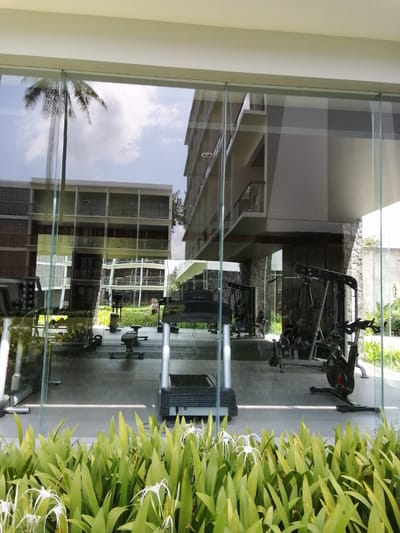 This image captures a reflection of an outdoor scene in the glass facade of a building, likely a hotel or resort, in Banyuwangi, Indonesia. The foreground is dominated by lush green foliage with delicate white flowers. Behind this, the glass reflects a gym area, featuring exercise equipment such as treadmills, stationary bikes, and weight machines. In the background of the reflection, a modern building with multiple stories, possibly apartments or hotel rooms with balconies, is visible. A palm tree is also reflected in the glass, hinting at the tropical location. The overall impression is one of a modern, well-maintained recreational facility integrated with its natural surroundings.