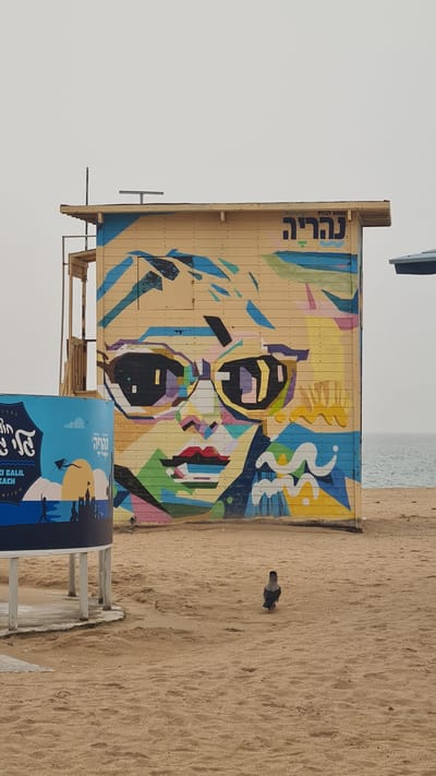 The image shows a beach scene in Nahariah, Israel. A lifeguard station stands on the sand, with a colorful mural of a face painted on its side. The mural is stylized and abstract, using geometric shapes and bold colors. Next to the station, there's a round advertisement or sign with lettering in Hebrew and English. In the background, the sea is visible, and a bird stands on the sand next to the station. The sky is overcast.