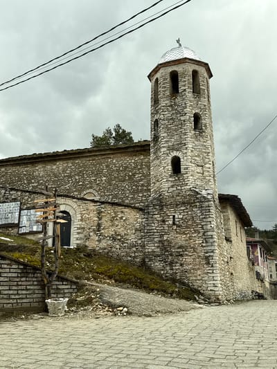 The image shows the Church of St. Theodore Tyron and Theodore Stratelates, located in Lilkovo, Bulgaria. The stone building consists of a tall tower with arched windows topped with a metal dome, and a lower section. A doorway is visible on the lower section, with a wooden structure placed in front of it. A paved walkway is to the right. Above the building are power lines against a cloudy sky.