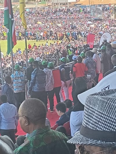 This image captures a large-scale outdoor public gathering or rally in Siaya, Kenya, under clear, bright daylight. The scene is densely populated with a vast crowd filling a stadium or expansive open field, extending far into the background.
In the foreground, on the left, a Kenyan flag (featuring black, red, and green stripes with a Maasai shield) is prominently displayed alongside a yellow flag. On the right, the backs of several individuals are visible. One prominent figure has a bald head and wears eyeglasses, dressed in a green garment with circular patterns. Nearby, another person wears a distinctive black and white checkered jacket. A white shirt or banner featuring the "Reebok" logo is also visible in the upper right.
The midground features a red-carpeted area or platform where key individuals are gathered. Many in this area are wearing matching green caps or hats and some are in red shirts, suggesting a unified group or supporters. Others are seen in light blue shirts. Behind this platform, a red banner with the white text "DTB" is clearly visible, along with a white sign bearing a unique logo (resembling a snowflake or star).
The sheer number of people in the background, tightly packed into the stands and across the field, indicates a significant event. Some members of the crowd are holding umbrellas, likely for sun protection. The overall atmosphere suggests a major public event, possibly a political rally, community celebration, or public address, characterized by widespread participation.