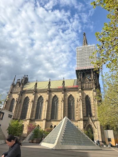 A vibrant daylight photograph captures a historic Gothic cathedral in Basel, Switzerland, framed by lush green tree canopies against a partly cloudy blue sky. The image is taken from a low angle, emphasizing the towering scale of the cathedral and its intricate stone architecture.
The prominent Gothic building features numerous pointed arches, large multi-paned windows, and decorative buttresses. Its roof is a distinct green hue. A significant portion of one of its towers is enveloped in white scaffolding, indicating ongoing renovation or maintenance. Visible through the scaffolding on the main facade is an ornate blue and gold clock face, adorned with Roman numerals.
Adjacent to the ancient structure, an angular, modern building with a distinctive textured glass block facade rises, creating a striking architectural contrast. This modern structure appears to be an entrance or skylight for an underground area. Parked bicycles are visible nearby, alongside some wooden crates.
In the foreground, partially visible in the bottom right, is a person with dark hair, looking downwards, adding a human element to the urban scene. Lush green trees with characteristic round seed pods and branches with light green leaves arch into the frame from the top, while flowering plants with purple blossoms add color near the modern building.
Below the cathedral facade, near the modern structure, a white sign clearly displays the text 'THEATER BASEL'. The overall impression is a bustling urban environment where history and modernity coexist under a bright, dynamic sky.