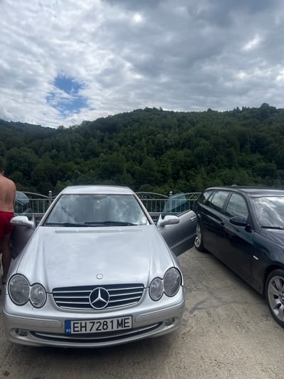 Two cars, a silver Mercedes-Benz CLK and a dark BMW 3 Series Touring, are parked on a paved surface overlooking a dense, green forest that covers a hillside. The sky is overcast with dramatic clouds. The Mercedes-Benz is in the foreground, its front end facing the camera, and its license plate reads "BG EH 7281 ME". The BMW is parked to the right of the Mercedes. A person, only their lower back and legs are visible, wearing red shorts, is standing to the left of the Mercedes, appearing to interact with the car. The setting appears to be outdoors, possibly in a natural or semi-natural environment given the prominent forest backdrop. This scene is located in Shipkovo, Bulgaria.
