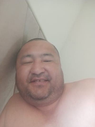 A selfie of a man is shown. He has light skin, a shaved head, and is smiling with his mouth open, revealing his teeth. The man is wearing a light-colored shirt and is looking at the camera. The background of the image is a light-colored wall. The image appears to be taken in a home environment in Mang'it, Uzbekistan.