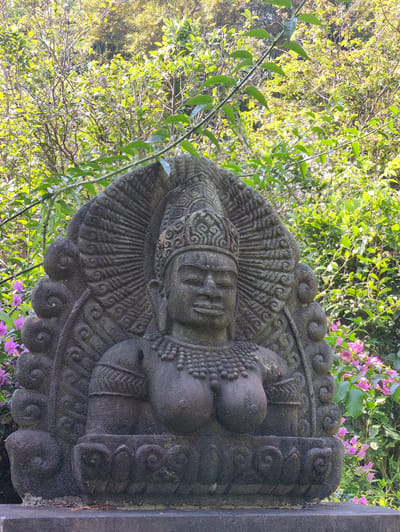 A stone sculpture rests in an outdoor setting, likely a garden or park. The sculpture appears to be a deity, perhaps a Buddha or Hindu figure, depicted with elaborate headdress, necklace, and ornamentation. The sculpture is made of dark, rough-hewn stone. The background features lush foliage, including green bushes and pink flowering plants. There is no evidence that this photograph was taken in Tuy Hòa, Vietnam.