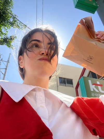 The image is a low-angle shot of a woman holding a brown paper bag. The bag obscures part of the view. A green sign with Korean text is partially visible above and to the side of the bag. The woman has dark hair and is wearing a white shirt with a red sweater over it. Sunlight is shining from behind the woman, creating lens flares and highlighting her features. In the background, there are trees, power lines, and buildings. The website on the bag, www.saeropni.com, indicates the location is likely in Daegu, South Korea. The Korean text "대구 식대" on the green sign suggests this is related to food or dining in Daegu.