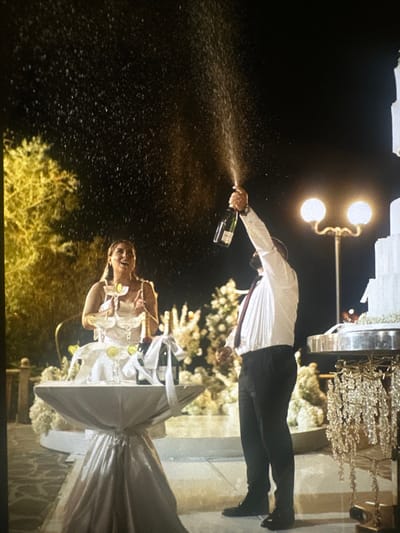 This image captures an outdoor nighttime event in Stains, France. A man, positioned on the right, wears a white long-sleeved shirt, a dark tie, and dark trousers. He holds a champagne bottle, identified as Moët & Chandon, with his right arm raised, spraying liquid upwards and to the left. To his left, a woman in a light-colored, possibly white, sleeveless dress stands smiling and looking upwards, appearing to react to the spraying champagne. Between the two individuals, a circular table draped in a light, reflective fabric (likely silver or white) holds several items. On this table, multiple champagne coupes are arranged in a tiered formation, each garnished with a lime wedge. Two additional champagne bottles, wrapped with white ribbons, are also present on the table. To the far right, a multi-tiered white cake on a silver stand, featuring ornate crystal-like dangling decorations, is partially visible. The background reveals illuminated trees to the left and a double-globe street lamp behind the man. The ground appears to be a paved surface. The sky is dark, indicating a nocturnal setting, with artificial lighting illuminating the scene.