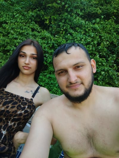 The media file shows a selfie of two people in front of a lush green hedge. The person in the foreground is a man with dark hair and a beard, looking directly at the camera with a slight smile. He is shirtless, and his skin appears damp, suggesting he may have been swimming or is in a humid environment. Behind him, and slightly to his left, is a woman. She has long dark hair and is wearing a leopard-print top. Her gaze is directed slightly away from the camera, and she also has a subtle expression. A tattoo is visible on her upper chest.
The setting is outdoors, with the dense green foliage of the hedge forming the backdrop for both individuals. The lighting suggests it is daytime, possibly late afternoon or early evening, with soft natural light. The presence of water droplets on the man's skin and the casual attire of both subjects hint at a leisurely activity, such as being by a pool or enjoying a warm day outdoors. There is no visible text in the image. The overall mood is relaxed and intimate.