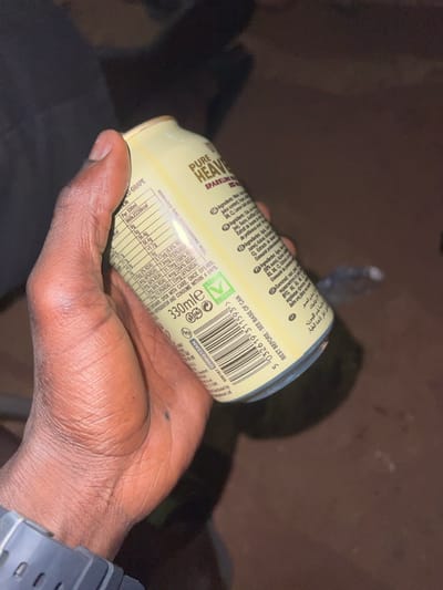 An individual's right hand, with dark skin tone, is holding a 330ml "PURE HEAVEN" sparkling drink can in a low-light environment in Birnin Kebbi, Nigeria. The hand grips the light yellow/cream-colored aluminum can, displaying its nutritional facts, ingredients in multiple languages (including English), barcode "5 032619 311593", and a green 'V' symbol. Visible text includes "Water, Fruit Juice from Concentrate (Grape 12%), Carbon Dioxide, Acid: Citric Acid, Natural Flavourings, Antioxidant: Ascorbic Acid, Vitamins: Niacin (B3), B6, B12, Folic Acid (B9), Biotin (B7)." A small, dark mark is present on the palm of the hand. The individual wears a dark grey digital wristwatch on the left wrist. The can's base exhibits a blueish discoloration and slight damage. The background consists of an indistinct, dark, reddish-brown ground surface, possibly soil or dirt, with blurred dark objects, including what appears to be a discarded plastic bottle, suggesting an outdoor or unpaved indoor setting.