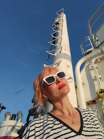 A woman with pink hair and white sunglasses poses for a selfie on a boat, with a blue sky and the mast of the boat in the background. She is wearing a striped shirt and a necklace with a crown pendant. The scene is set in the city of Shchetinino, Russia.