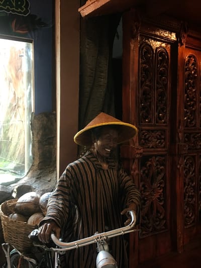 A man wearing a striped shirt and a conical hat stands next to a bicycle, smiling. He is in front of a set of intricately carved wooden doors.  A basket of coconuts is in the foreground. The scene appears to be in the Batu Secret Zoo in Batu, East Java, Indonesia.  The coordinates -7.884, 112.525 place this location near the entrance of the zoo.