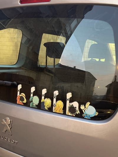 A silver Peugeot car is parked outdoors, reflecting the sky and buildings. The rear window of the car features a sticker of several cartoon characters from the anime series One Piece, with their arms raised in a unified gesture. The characters, from left to right, appear to be Monkey D. Luffy, Roronoa Zoro, Vinsmoke Sanji, Nami, and Tony Tony Chopper. The stickers are placed along the bottom edge of the rear window. In the background, a streetlamp is visible, illuminating a street with buildings on either side. The car's rear bumper is visible at the bottom of the image, with the Peugeot logo to the left. The scene is likely set in a residential area in Santa Coloma de Gramenet, Spain, given the architectural style of the buildings and the streetlamp.