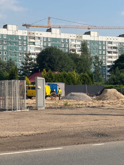 The image depicts a construction site adjacent to a large apartment building in Novoe Devyatkino, Russia. A tall, orange construction crane dominates the left side of the frame, its boom extending over the building. The apartment building itself is a multi-story structure with a distinctive light blue-green facade and white accents around the windows.
In the foreground, a dirt and gravel area forms part of the construction site. Several construction vehicles are visible, including a yellow van and a blue generator. A person wearing a high-visibility yellow vest is standing near the vehicles, possibly overseeing or participating in the work. A large pile of gravel and a substantial rock are also present on the site.
In front of the construction vehicles and a portion of the fence, there is a silver, rectangular structure with a mesh-like surface, possibly a piece of equipment or a temporary shelter. Further towards the bottom of the image, metal fencing or barriers are stacked, indicating ongoing construction or site preparation.
The scene appears to be set during the daytime, with clear skies suggesting fair weather. The presence of construction equipment and the partially visible apartment building suggests an ongoing building project or renovation. The overall impression is one of active development in a residential area. No discernible text is visible in the image.