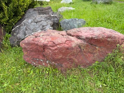 A red boulder rests in the grass in Akatovo, Russia. The boulder is roughly spherical and appears to be made of sandstone. It is surrounded by green grass and a few small white flowers. In the background, there are more boulders and a large green bush. The sky is not visible in the image.