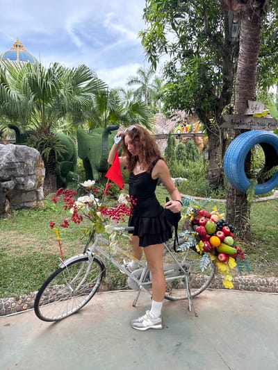 A woman with curly brown hair, wearing a black dress and white sneakers, stands with one foot on the pedal of a white bicycle. The bicycle's front basket is overflowing with a colorful arrangement of fruits and flowers, including apples, oranges, grapes, and red berries. A red flag is affixed to the bicycle's handlebars. In the background, green, topiary-like elephant sculptures are visible, along with palm trees and a large building with a blue dome. A blue tire is mounted on a tree to the right. The scene appears to be an outdoor park or garden.