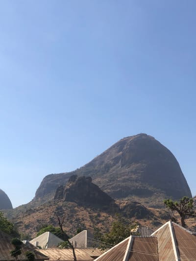 The image displays a landscape in Bundot, Nigeria, under a clear, cloudless blue sky. A large, prominent, rounded rocky mountain with sparse, dry, brown vegetation dominates the mid-ground, situated slightly right of center. To its left, a smaller, darker, and more jagged rock formation rises from the terrain, also featuring dry vegetation. In the foreground, the roofs of multiple buildings are visible, constructed from peaked or gabled corrugated metal sheets, predominantly showing rust-brown tones. Sparse trees with limited green foliage are scattered in the lower areas of the scene, notably a distinct tree with a thick trunk and sparse branches on the far right. No individuals, specific actions, or interactions are present in the scene.