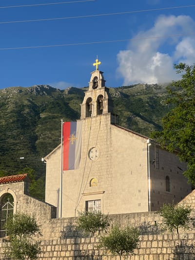 The image displays a serene outdoor scene featuring a stone church building against a backdrop of mountainous terrain and a clear blue sky.
In the center of the frame is a light-colored stone church with a prominent bell tower. The bell tower is topped with a gleaming golden cross, reflecting bright sunlight. Below the cross, the tower features arched openings where bells are visible. Attached to the facade of the church building, below the bell tower, is a flag. The flag is a horizontal tricolor of red, blue, and white. In the center of the blue stripe, there is a golden emblem, which appears to be a cross with four C-shaped elements (a common symbol associated with the Serbian Orthodox Church). The church building itself is constructed from cut stone blocks and features circular decorative elements on its facade and a window with a simple grid pattern.
Behind the church, a large, rugged mountain range is visible, densely covered with green trees and vegetation. Patches of white clouds partially obscure the mountain peaks. The sky above is a vibrant blue, with a few scattered white clouds. Several thin, dark power lines stretch horizontally across the upper portion of the sky.
In the foreground and to the sides of the church, there are small green trees or bushes, and the ground or surrounding structures are paved with similar light-colored stone, some of which shows subtle shadow patterns.
The scene depicts a peaceful daytime setting, likely under bright sun, given the strong illumination on the church's cross and facade. There are no people visible, and no specific activity is taking place. There is no discernible text in the image. The overall impression is one of natural beauty and architectural heritage in a mountainous region, consistent with the Pelinovo, Montenegro context.