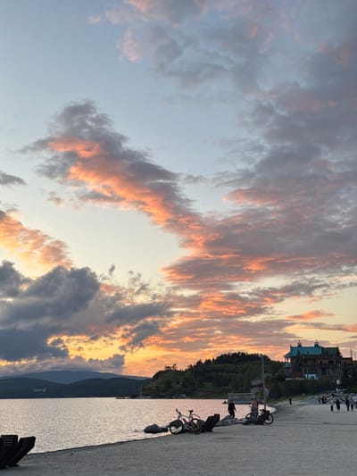 This is a wide shot of a beach in Turgoyak, Russia, at sunset. The sky is filled with dramatic clouds, tinged with orange and pink from the setting sun. A calm body of water, likely Lake Turgoyak, stretches across the left side of the image, with a dark mountain range in the distance. The beach itself is sandy, with several beach chairs, two bicycles, and scattered rocks. On the right side of the beach, there are several people walking and standing, and a large, multi-story building with a teal roof is visible on a wooded hill overlooking the scene.