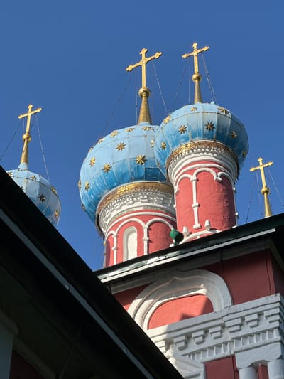 The image shows a close-up, low-angle view of the upper portions of multiple onion domes of a Russian Orthodox church, set against a clear, bright blue sky. The domes are predominantly blue with gold stars, topped with gold crosses. The lower sections of the domes are red with white decorative trim. A dark, angled roofline, possibly of another building or an overhang, partially obscures the bottom left of the frame. The architectural style suggests a traditional Russian church. The city of Terpilovo, Russia, is mentioned as the location. There are no people visible in the image.