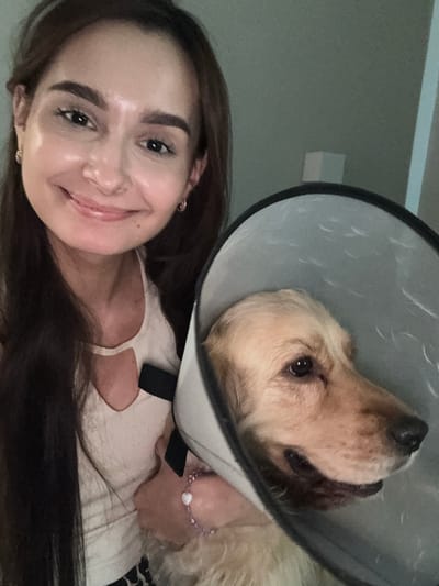 A woman with long, dark hair and a light-colored tank top is taking a selfie with her golden retriever dog, who is wearing a protective cone. The woman is smiling and looking at the camera. The dog is looking to the right, with its mouth slightly open. The background is a plain wall with a subtle shadow. The location is identified as the city of Kuyuki, Russia.