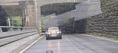 An orange and blue vintage-style sports car with a license plate reading "GF 69 CE" drives through a tunnel in Golling an der Salzach, Austria. The car is in the center of the frame, moving away from the viewer. The road is wet, indicating it may be raining. The tunnel walls are made of concrete and stone. A yellow and red striped pole is visible to the left of the car. Traffic lights and speed limit signs are also present. The overall environment appears to be a road leading into or out of a tunnel, possibly in a mountainous region given the stone wall.