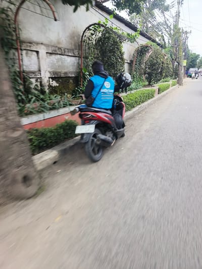 The scene depicts a roadside in Cimahi, Indonesia, under overcast daylight. A person, observed from behind, is seated on a red scooter positioned at the edge of an asphalt road, facing away from the viewer. The individual wears a blue vest with the "inDriver" logo over a dark long-sleeved garment and a dark head covering. A black motorcycle helmet is visible, suspended from the scooter, and a black bag or object rests on the scooter's floorboard near the rider's left foot. The scooter's rear features a blurred white license plate.

To the left of the scooter, a low concrete curb separates the road from a landscaped strip containing a manicured line of green hedges. Behind these hedges, a light-colored concrete wall extends horizontally, detailed with recessed rectangular sections and multiple metal arch-shaped trellises supporting climbing green plants. A large tree trunk partially obstructs the immediate foreground on the left. Further down the road, other motorbikes and a light blue/green vehicle are visible in the distance, indicating traffic. Utility poles and additional trees line the far side of the street.