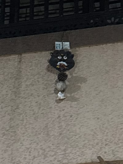 The scene is set against a textured, light-brown building wall in Ambernath, India, captured under low light. A decorative hanging item is centrally affixed to this wall. The item comprises a dark-colored, stylized head-like form featuring white, circular eyes with dark pupils, a white nose, and an open mouth detailed with white, red, and yellow elements. Two light-colored, ear-like projections extend laterally from the head. Vertically below the head, the item incorporates a dark, spherical, textured component, followed by multiple lighter-colored, amorphous elements, and concludes with a small white object at its base. A thin filament suspends the item from a small, light-colored rectangular tab secured to the wall directly above it. A dark, horizontal architectural structure, consistent with a railing or ledge, occupies the upper background. No individuals or specific actions are depicted.