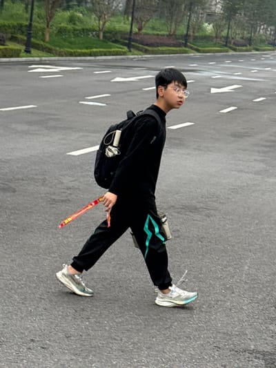 An outdoor, full-body shot captures a young individual standing on a paved road in Shuangfu, China. The road is marked with white dashed lines, and to the left, a lush green landscape of trees and foliage forms the backdrop.

The subject is a young person, possibly male, with short dark hair and wearing clear-rimmed glasses. They are dressed in a casual all-black outfit, consisting of a long-sleeved top or jacket and track pants. The left pant leg features a distinctive teal or light blue zigzag pattern running along the side. They wear light-colored athletic sneakers, predominantly white with hints of blue. A black backpack is slung over their shoulders, with a white, cylindrical object (possibly a water bottle or thermos) attached to its side.

The person is captured looking upwards and slightly to their right, with a neutral and observant expression, their mouth slightly agape. In their right hand, they hold a long, red and yellow object, which appears to be a packaged snack or a novelty item like a firecracker stick, featuring some unidentifiable Chinese characters.

The scene appears to be set during daytime, with soft, even lighting suggesting either an overcast sky or early/late daylight hours. The road surface is dry. The overall impression is one of a calm moment of contemplation or observation within a semi-urban or park-like environment.