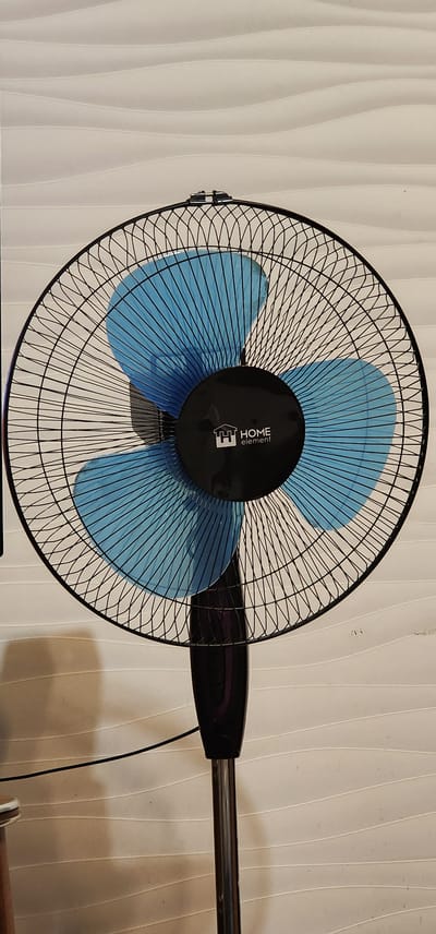 A black, oscillating stand fan with bright blue blades is positioned centrally in the frame against a textured, off-white wall. The fan's guard is made of thin black wires, creating a crisscross pattern. In the center of the fan, a black circular housing displays the white logo and text "HOME element." The fan is mounted on a black plastic stem that transitions to a chrome-plated pole supporting it. A thin black power cord extends from the fan's base and runs horizontally to the left. The background wall features subtle horizontal wavy lines. There are no people visible in the image. The image is a photograph taken in a room, possibly a residential or commercial space.
