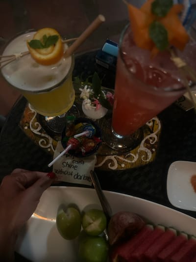 The image displays a high-angle view of a table setting within a dimly lit establishment, likely in Thuận Hóa Ward, Vietnam. Two garnished cocktails are positioned on a round, dark brown tray featuring decorative gold and white swirl patterns. The cocktail on the left is a yellow, frothy drink in a stemmed glass, garnished with a spiral orange peel and mint leaves, accompanied by a bamboo straw and two wooden skewers. The cocktail on the right is a red drink with ice, served in a tall stemmed glass, garnished with a piece of carved orange-colored fruit (possibly starfruit or papaya) and mint leaves, also with a bamboo straw.
Between the cocktails, on the decorative tray, rests a small white vase containing small white and pink flowers with green foliage. Adjacent to the flowers, a small blue bowl holds an assortment of colorful lollipops and candies, some with visible wrappers (one reading "MELLY").
Below the tray, a white rectangular dish contains an arrangement of cut fruits: several green plums or limes (some scored), multiple slices of watermelon, and slices of a reddish fruit, possibly apple or plum. A metallic utensil, appearing to be a spoon, is visible beside the fruit. To the right of the fruit dish, a small white square dish contains a reddish-orange powder, likely a spice mix or chili salt.
A person's hand, with red painted fingernails, is visible in the lower-left foreground, holding a small, light-colored note. The handwritten text on the note reads: "Stars can't Shine without darkness." In the background, partially obscured, is a blue object, possibly a cigarette pack, with some text visible. The table surface beneath the items is dark with a textured or patterned fabric.