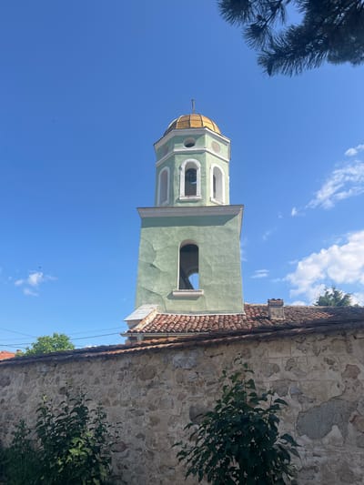 The image shows a bell tower in Zelenikovo, Bulgaria, on a clear, sunny day. The bell tower is painted light green with white trim and has a golden dome. It is situated behind a stone wall and a tiled roof. Green foliage is visible in the foreground and on the left side of the image. A few small clouds dot the bright blue sky, and a bird is visible in flight. The architecture suggests a historical or religious building.