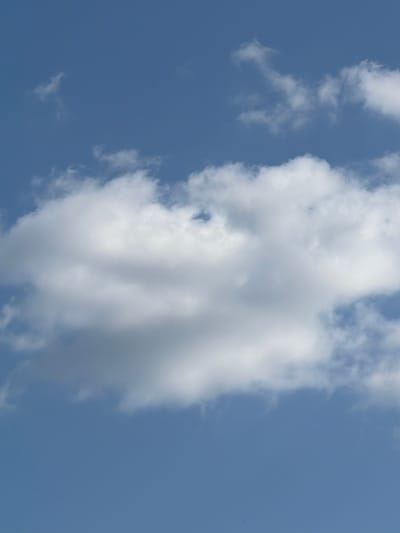The image shows a clear blue sky with several white clouds. The largest cloud is in the center of the frame and is puffy and textured. There are smaller, more wispy clouds scattered across the sky. The lighting suggests it is daytime, possibly mid-morning or afternoon. The image was taken outdoors and does not show any buildings or ground. It's a typical sky scene with clouds.
